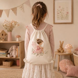 May include: A cream-colored drawstring backpack featuring a printed illustration of a fairy with pink wings and a pink dress, sitting among flowers. Below the fairy, the name Mila is written in a cursive black script font on the fabric.