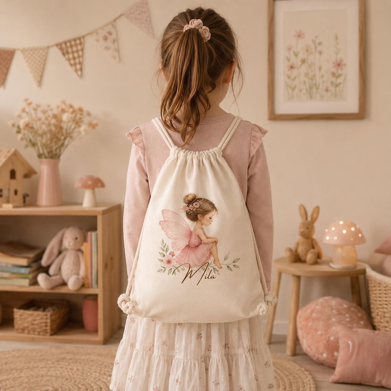 May include: A cream-colored drawstring backpack featuring a printed illustration of a fairy with pink wings and a pink dress, sitting among flowers. Below the fairy, the name Mila is written in a cursive black script font on the fabric.