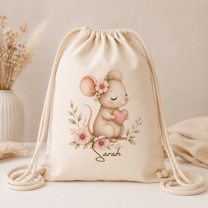 May include: A cream-colored drawstring backpack featuring a printed illustration of a small mouse with pink ears, holding a pink heart, surrounded by pink flowers and green leaves. The name Sarah is printed in brown cursive script below the mouse.