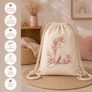 May include: A natural-colored drawstring backpack featuring a printed illustration of a white unicorn with a pink mane, floral accents, and the name Aurelia in cursive script. The bag has thick cream-colored rope straps and a woven fabric texture.