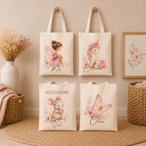 May include: Four off-white canvas tote bags featuring illustrations of a fairy, a unicorn, a mouse holding a heart, and a butterfly. The bags include the printed names Mila, Aurelia, Sarah, and Olivia, and the text WECHSELKLEIDUNG on one bag.