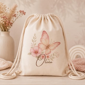 May include: A cream-colored drawstring backpack made of canvas fabric. The front features a printed design of a large pink butterfly, a pink flower, and green leaves. The name Olivia is printed in a dark brown cursive script below the butterfly.