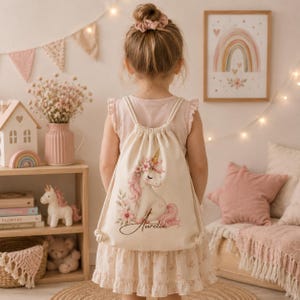 May include: A cream-colored drawstring backpack featuring a printed illustration of a white unicorn with a pink mane, floral accents, and the name Aurelia written in cursive script. The bag has matching cream-colored rope straps.