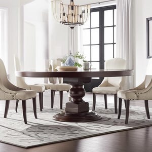 Può includere: Un tavolo da pranzo rotondo, marrone scuro, con base a piedistallo è circondato da sei sedie imbottite beige. Le sedie hanno gambe in legno scuro e sono disposte su un tappeto fantasia. Un lampadario è appeso sopra il tavolo.