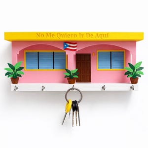 May include: A pink and yellow key holder shaped like a house, with the text "No Me Quiero Ir De Aqui" above the roofline. It features a Puerto Rican flag, two windows, a door, and key hooks.