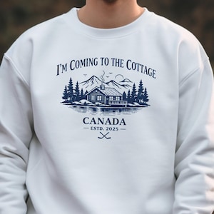 Może przedstawiać: Biała bluza z granatową grafiką z napisem "I'M COMING TO THE COTTAGE" nad sceną z domkiem, słowem "CANADA" i "ESTD. 2025". Projekt zawiera drzewa, góry i skrzyżowane kije hokejowe.