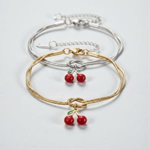 Peut inclure: Deux bracelets fins, un argenté et un doré, sont présentés. Le bracelet doré est orné d'un charm cerise avec des fruits rouges et des feuilles vertes. Le bracelet argenté est au-dessus du bracelet doré. Les deux bracelets ont des fermoirs réglables.