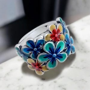 Può includere: Un anello d'argento con un design floreale. L'anello presenta fiori smaltati blu, verdi, gialli e rossi. L'immagine è un primo piano, che evidenzia gli intricati dettagli dell'anello e i colori vivaci dei fiori.