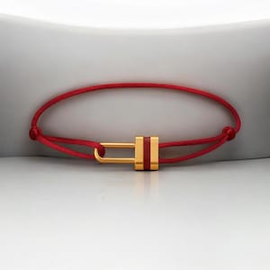 May include: A red cord bracelet with a gold-coloured rectangular clasp. The clasp has a gold-coloured frame with a red stripe in the centre. The bracelet is displayed on a white surface.