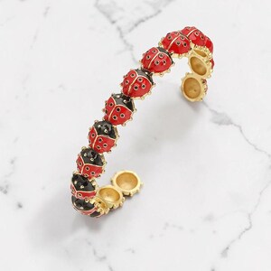 May include: A gold-toned bracelet featuring a series of red and black ladybug charms. The ladybugs are detailed with black spots and are connected by gold links. The bracelet is set against a white marble background.