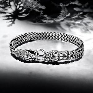 May include: A silver-toned bracelet with a snake design. The bracelet features a woven chain and two snake head clasps that connect with a circular clasp. The bracelet is displayed on a reflective surface.