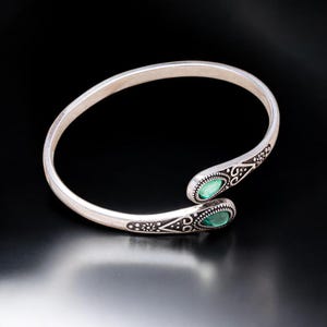 Puede incluir: Brazalete de puño plateado con diseño abierto. El brazalete presenta dos piedras preciosas verdes de forma ovalada en los extremos. Intrincados patrones oscuros grabados adornan los terminales del brazalete, añadiendo un toque decorativo.