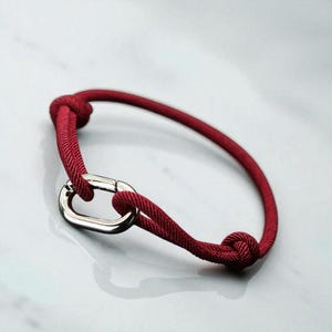 May include: A red rope bracelet with a silver clasp. The bracelet is made of a thick, woven rope material and has a knot at each end. The clasp is a simple, oval design.