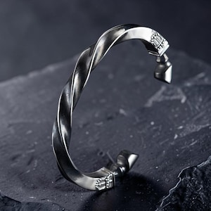 May include: A silver-colored, twisted metal bracelet. The bracelet has a textured, rope-like design and decorative end pieces. The ends feature a small, ornate design. The bracelet is displayed on a dark, textured surface.