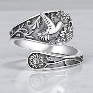 May include: A silver-colored wrap ring featuring a hummingbird design. The ring has a detailed floral and hummingbird motif, with sunflowers and leaves. The ring wraps around the finger, creating a unique and elegant look.