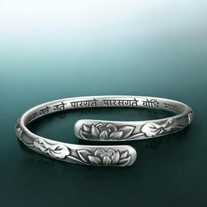 May include: A silver-colored cuff bracelet with a floral lotus design and engraved text. The open-ended bracelet features detailed lotus flower carvings and a Sanskrit inscription. The bracelet is displayed on a dark teal background.
