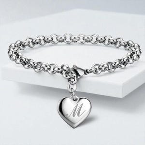 Könnte beinhalten: Ein silberfarbenes Armband mit einem Kettenglied-Design. Ein herzförmiger Anhänger mit dem eingravierten Buchstaben "M" hängt an dem Armband. Das Armband wird auf einer weißen Oberfläche präsentiert.