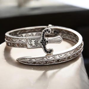 Puede incluir: Brazalete plateado con diseño de espada. El brazalete presenta intrincados adornos y una espada con detalles de piedras preciosas negras. La espada está colocada sobre la abertura del brazalete, creando un diseño único.