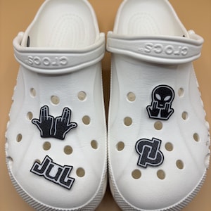 Puede incluir: Un par de zuecos blancos sin cordones con adornos decorativos en blanco y negro. Los adornos incluyen un gesto con la mano, el texto "JUL", una cabeza de alienígena y un logotipo geométrico. Los zuecos tienen una tira pivotante en el talón.