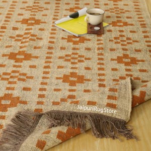 May include: Beige and orange patterned rug with a geometric design. The rug has a fringed edge and is partially rolled up, revealing the underside. A cup of coffee, a coaster, and a pen are on top of a yellow notebook.