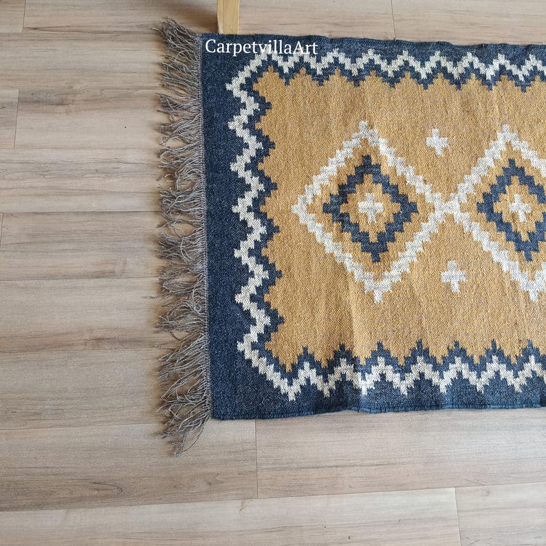 May include: A rectangular rug with a geometric pattern. The rug features a mustard yellow center with diamond and cross designs, framed by a navy blue border with a white zigzag pattern. The rug has a fringe on one end.