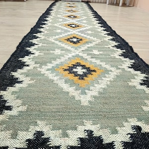 Puede incluir: Una alfombra larga y rectangular con un patrón geométrico. La alfombra presenta un diseño central en tonos grises, blancos y amarillos, enmarcado por un borde negro. El texto "CarpetvillaArt" es visible en la esquina inferior izquierda.