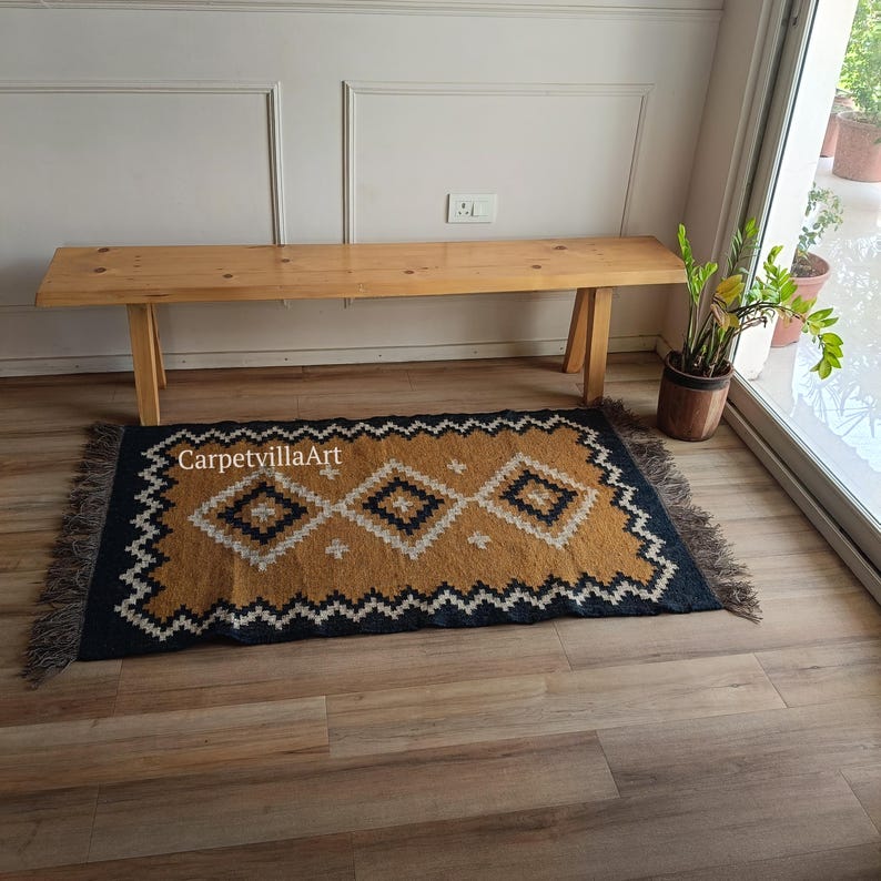 May include: A rectangular rug with a geometric pattern in mustard yellow, navy blue, and white. The rug has a fringed edge and the text "CarpetvillaArt" is visible. The rug is placed on a wooden floor.