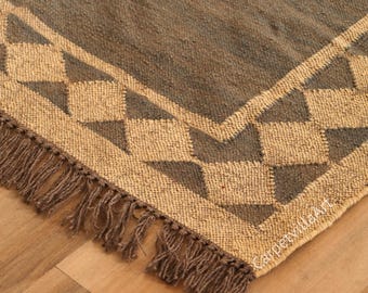 May include: A close-up of a brown and tan jute rug with a geometric diamond pattern border. The rug has a fringed edge and is made of natural fibers. The rug is a decorative home accent.