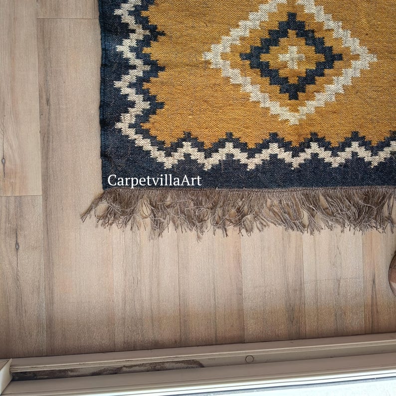 May include: A rectangular rug with a geometric diamond pattern in mustard yellow, navy blue, and white. The rug has a fringed edge and the text "CarpetvillaArt" is visible. The rug is placed on a light wood floor.