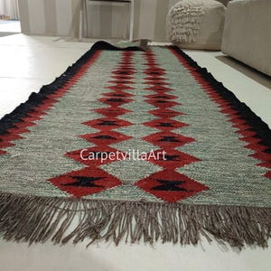 Può includere: Un tappeto rettangolare con un motivo geometrico. Il tappeto presenta uno sfondo grigio con un motivo a rombi ripetuto in rosso e nero. I bordi sono neri e il tappeto ha una frangia alle estremità. Il testo "CarpetvillaArt" è visibile sul tappeto.
