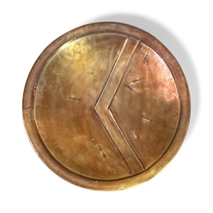 May include: A round, bronze-colored shield with a prominent chevron design. The shield has a slightly weathered appearance, suggesting an antique or historical aesthetic. The shield is set against a white background.