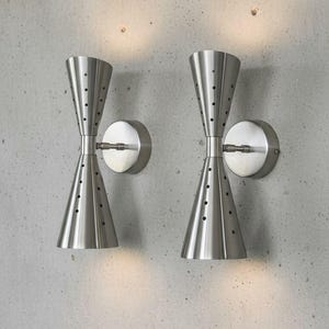 Puede incluir: Dos apliques de pared de níquel cepillado con un diseño moderno de mediados de siglo. Cada lámpara tiene forma de reloj de arena con pequeñas perforaciones y una placa de montaje circular. Los apliques están montados en una pared de hormigón gris.