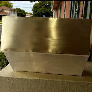 May include: A rectangular planter box with a gold-colored top and a white, trapezoidal base. The gold top has a brushed metal appearance, reflecting light. The planter is designed for outdoor use and is made of durable materials.