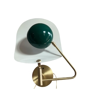 Peut inclure: Une applique murale moderne avec un abat-jour sphérique vert foncé et un réflecteur blanc incurvé. La lampe est dotée d'un bras et d'une base de couleur laiton, conçue pour l'éclairage intérieur. Le design est minimaliste et élégant.