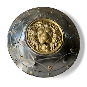 May include: A round, silver-colored shield with a gold-colored lion's head in the center. The shield has a series of rivets and decorative elements. The lion's head is detailed with a mane and facial features.