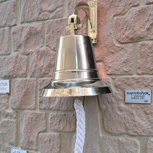 May include: A polished brass ship's bell mounted on a brick wall. The bell has a flared base and a white rope for ringing. The mounting bracket is also brass. The wall is made of reddish-brown bricks.