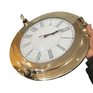Könnte beinhalten: Eine runde Uhr in Messingfarbe mit weißem Zifferblatt und schwarzen römischen Ziffern. Die Uhr hat ein nautisches Bullauge-Design mit einem Klappverschluss und einem Griff. Die Zeiger der Uhr sind schwarz mit einem roten Sekundenzeiger.