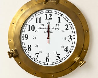May include: A round wall clock with a brass porthole-style frame featuring a white face, black Arabic numerals for 1-12 and 13-24 hour markers, and the text Edmund Asnley LONDON. The clock has black hour and minute hands and a red second hand.
