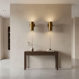 May include: Two gold wall sconces with cylindrical shades light up a neutral wall. A dark wood console table below holds a small vase with dried flowers and a stack of books. The room has a minimalist design with a tiled floor.