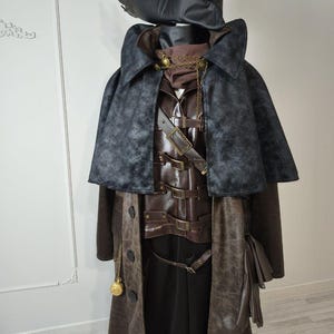 May include: A detailed costume featuring a black leather hat, a dark gray cape, and a brown leather vest with multiple buckles. A long brown coat with a pocket and a gold pocket watch completes the ensemble. The costume is designed with a vintage aesthetic.