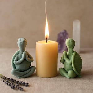 Puede incluir: Una vela encendida de color crema con dos figuras de tortugas en postura de yoga. Una tortuga es verde salvia, la otra es verde azulado. Una ramita de lavanda está en primer plano. Cristales en el fondo.