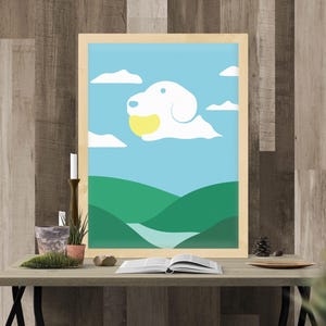Könnte beinhalten: Ein gerahmter Kunstdruck mit einer skurrilen Illustration einer hundeförmigen Wolke, die einen gelben Ball vor blauem Himmel hält. Das Kunstwerk befindet sich über grünen Hügeln und einem Fluss. Der Druck wird auf einem Holztisch mit dekorativen Gegenständen ausgestellt.