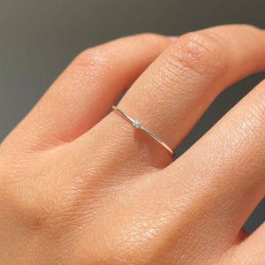 May include: A delicate silver ring with a small, clear gemstone. The ring is thin and minimalist, featuring a single, sparkling accent. The design is simple and elegant, suitable for everyday wear or special occasions. The ring is shown on a hand.