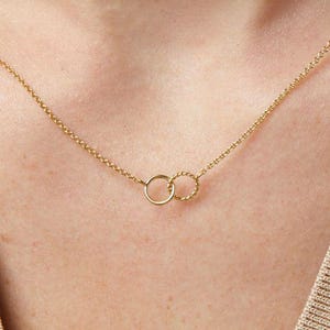 May include: A delicate gold necklace featuring two interlocking circles. One circle is smooth, while the other has a textured, twisted design. The necklace is displayed on a person's skin, highlighting the jewelry's elegant simplicity.
