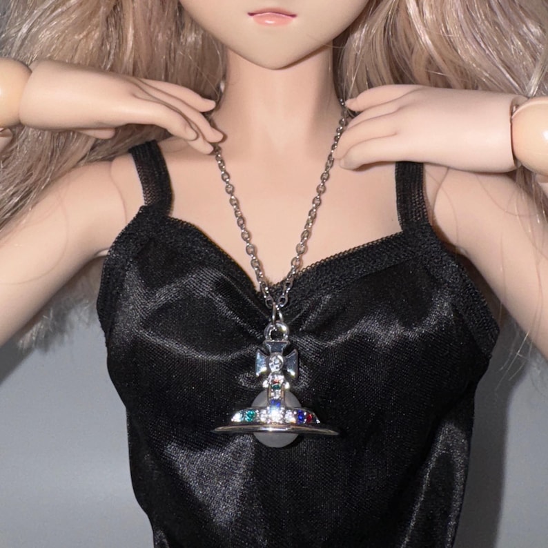 Puede incluir: Un collar plateado con un colgante de planeta con una cruz y gemas de colores. El collar se lleva sobre un top de sat&eacute;n negro. Se ven las manos de la mu&ntilde;eca.