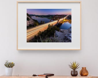 May include: A framed landscape photograph featuring a long-exposure view of a bridge over a river at twilight. The image captures light trails from traffic moving across the bridge, with rocky cliffs, dark trees, and a gradient sky at dusk.