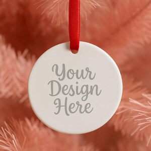 Puede incluir: Una bola de Navidad blanca y redonda con el texto "Your Design Here" en escritura gris. El adorno cuelga de una cinta roja sobre un fondo rosa suave, perfecto para decoraciones navideñas personalizadas.
