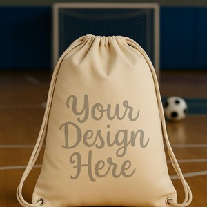 Könnte beinhalten: Ein beigefarbener Turnbeutel mit dem Text "Your Design Here" in grauer Schrift. Die Tasche hat einen Kordelzugverschluss und steht auf einem Holzboden. Im Hintergrund ist ein Fußball zu sehen.