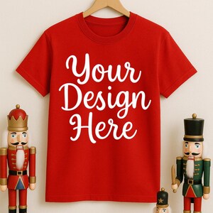 May include: A red t-shirt with the text "Your Design Here" in white script. The shirt hangs on a wooden hanger. Two nutcracker figures stand on either side of the shirt, with a small gift box in front.