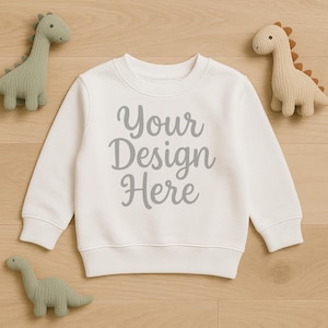 Puede incluir: Una sudadera blanca de cuello redondo con el texto "Your Design Here" en escritura gris. La sudadera está rodeada de tres juguetes de dinosaurios de peluche en colores verde y beige. Los artículos están colocados sobre una superficie de madera clara.